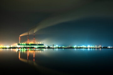 Naklejka na ściany i meble Brightly illuminated coal power plant high pipes with black smoke moving upwards polluting atmosphere at night with reflections of it in lake water.