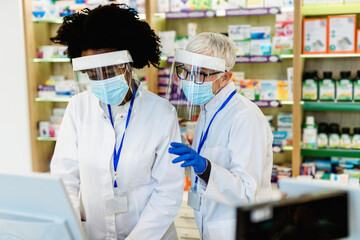 Black and Caucasian professional pharmacists working in modern drugstore. They are wearing face protective masks and shield for protection from virus disease. Coronavirus, Covid-19 concept.