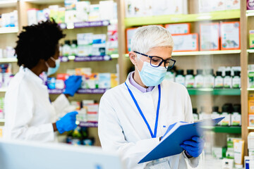Black and Caucasian professional pharmacists working in modern drugstore. They are wearing face protective masks and shield for protection from virus disease. Coronavirus, Covid-19 concept.
