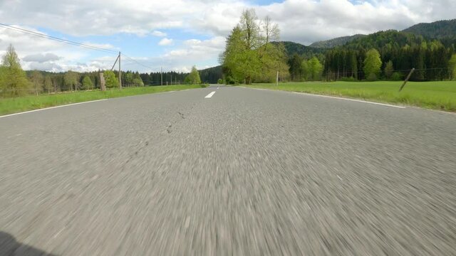 POV: Speeding down the empty asphalt road in the idyllic Slovenian countryside. Picturesque rural landscape surrounds the empty scenic route in Slovenia. Fast driving along a weathered country road.