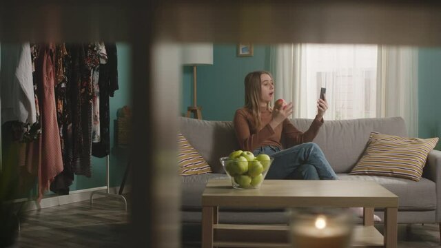 Pretty lady in jeans and brown sweater sits on beige sofa on window background, bites red apple and talks on video call. On coffee table is glass jar with green apples. Camera dollies horizontally