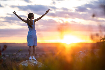 A young woman in summer dress raising up her hands standing outdoors enjoying view of bright yellow sunset.