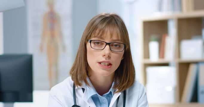 Close Up Portrait Of Young Beautiful Serious Female Caucasian Doctor In Glasses Sitting In Hospital And Speaking Looking At Camera Explaining Treatment, Telling Analysis Results, Healthcare Worker