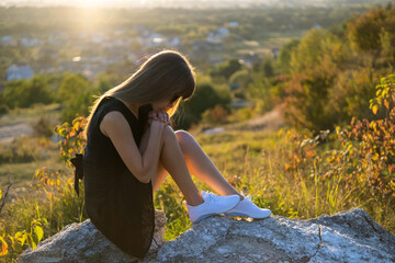 Young depressed woman in black short summer dress sitting on a mountain hill thinking outdoors at...