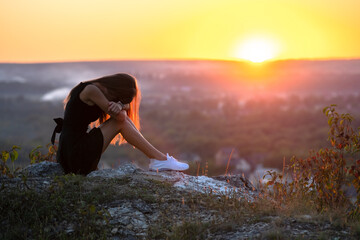 Young depressed woman in black short summer dress sitting on a rock thinking outdoors at sunset....