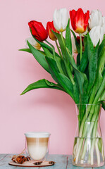 Beautiful bright bouquet of tulips in a transparent vase and a cup of cappuccino isolated on a pink background. Delicate minimalistic composition of spring flowers.