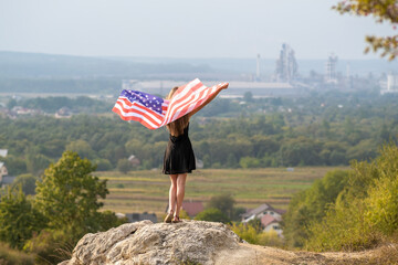 Obraz premium Young happy american woman with long hair raising up waving on wind USA national flag in her hands relaxing outdoors enjoying warm summer day.