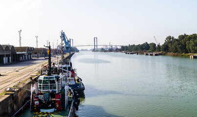 Two tugboats moored in the Guadalquivir river next to the warehouses and industrial units of the port of Seville on a sunny day (Andalusia, Spain). Views of the Centennial Bridge in the background.