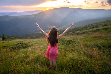 Rear view of a happy woman hiker in red dress standing on grassy hill on a windy evening in autumn mountains with outstretched arms enjoying view of nature at sunset.