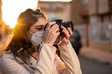 Joven mujer fotógrafa con una cámara réflex negra haciendo una foto al atardecer en un paisaje urbano, con cazadora blanca, camiseta de rayas, al atardecer