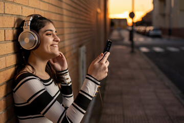 Mujer joven caucásica escuchando música con auriculares marrones y plateados con su smartphone...