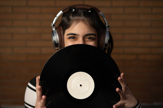 Mujer joven de pelo negro caucásica con auriculares y un disco de vinilo negro tapando su cara, enseñando sus ojos, sonriendo