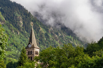 bell tower with a clock in a mountain village. Clouds cover a hillside in the background, in...