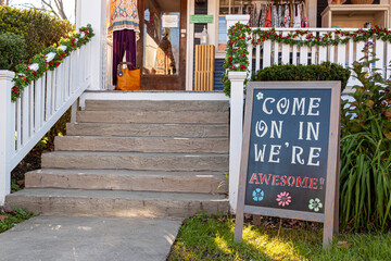 A sandwich board with a welcoming notice on it which reads : " Come on in, we are awesome " is placed in the front yard of a small local clothing shop in historic Clifton, Virginia.