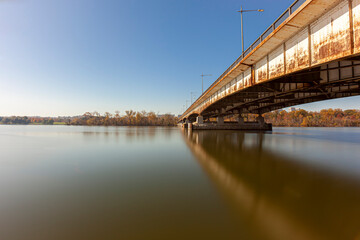 Long exposure landscape image featuring Theodore Roosevelt Island and bridge and Potomac river on a sunny afternoon in autumn. Trees on island have orange red brown colors. The bridge is rusty .