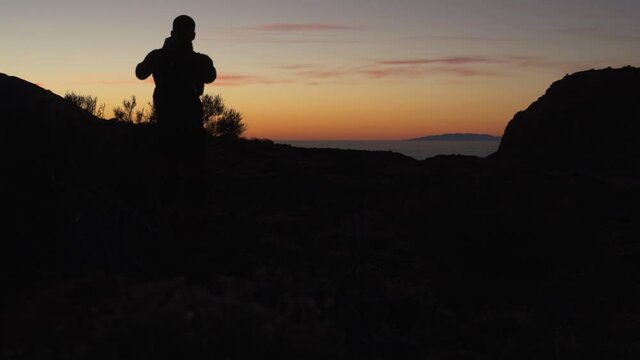 Silhouette of backpacker hiker taking picture of the beautiful after sunset orange sky over the Atlantic ocean in Tenerife island.