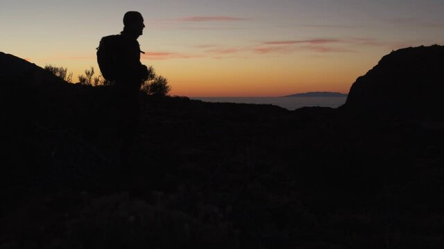 Silhouette of backpacker hiker taking a moment to capture picture of the beautiful after sunset orange sky over the Atlantic ocean in Tenerife island.