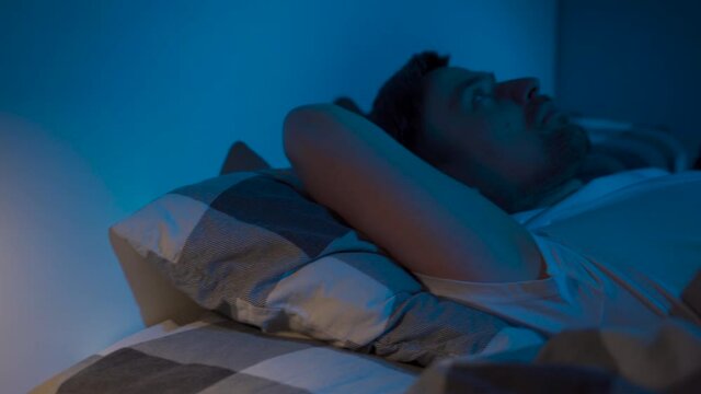 Depressed man lying in bed cannot sleep from insomnia. Young male trying to sleep. Restless worried guy wakes up at night lying on bed, sleepless, wide-eyed, suffering from insomnia, sleep disorder