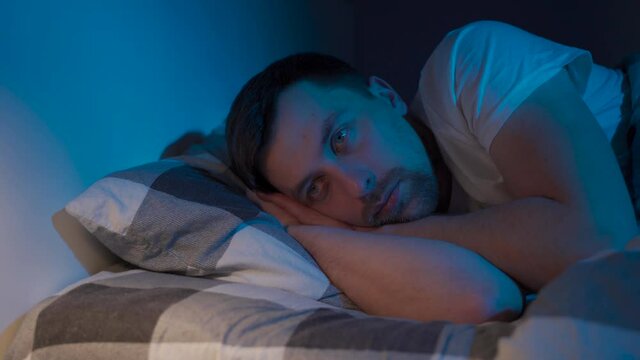 Stressed man trying to sleep in bed at night. Young male with open eyes suffers from insomnia and sleep disorders. Things dont feel same anymore. Blue tones. Sleepless man. Guy dont sleep at night