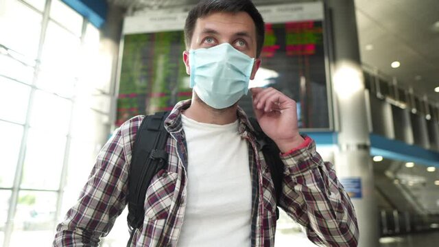 Coronavirus and travel on public transport. Caucasian man complies with new rules by wearing a protective medical mask in a public place at an airport or passing a train station near flight panel