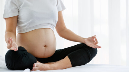 close up of a tummy of a healthy pregnant woman sitting on a white bed doing Yoga for excise in a bedroom at home. Selective focus on the belly of a lady