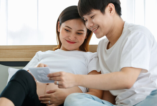 portrait of a very happy young Asian man and pregnant woman sitting on a bed smiling and looking at an ultrasound scan photo of fetus. Happy and healthy family concept