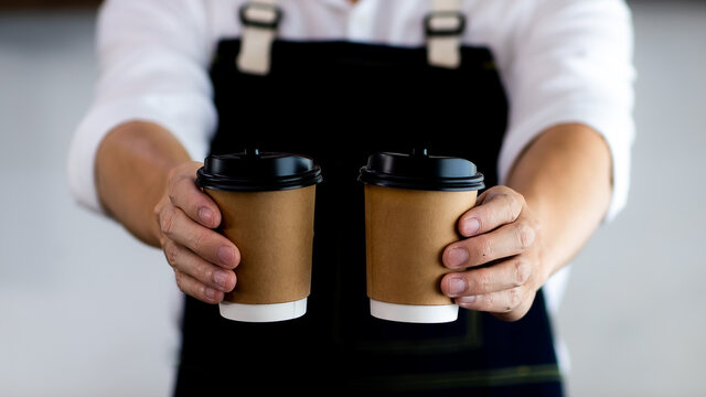 Asian Young Man Smiling Holding Paper Coffee Cup. At The Bartender In The Coffee Shop. Preparing For Pressing Ground Coffee For Brewing Espresso Or Americano In A Cafe. Concept Coffee Maker In Cafe.