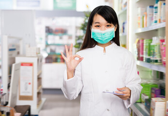 Adult chinese female in protective facial mask showing ok hand sign in pharmacy