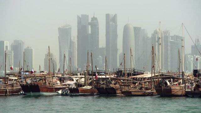 Large Cluster Of Wooden Dhow Boats Rocking Gently In Harbor With Fluttering Flags Before Stunning Cityscape