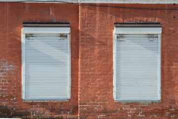Shutters. Old facade painted red brick wall with 2 windows closed by white roller shutters, illuminated by bright sunlight