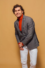 Obraz premium Tall handsome man dressed in orange turtleneck, white jeans and grey jacket posing on the beige background