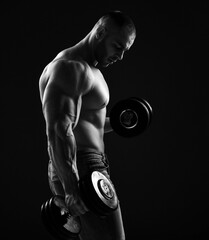 Muscular men bodybuilder is working out in gym, lifting alternately two dumbbells, doing exercises for biceps and looking down over black background. Young man lifting weights. Black and white  - 419538686