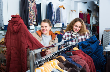 Two young cheerful women shopping in outerwear clothing boutique