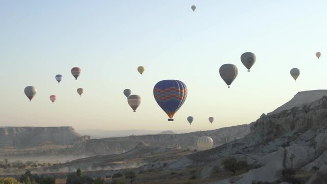 Various coloured tourist hot air balloons drift on different heights over bare rocky cliffs as festive parade under orange blue morning sky.