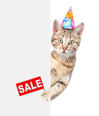 Cat wearing a birthday hat looks from behind empty white banner and holds sales symbol. isolated on white background