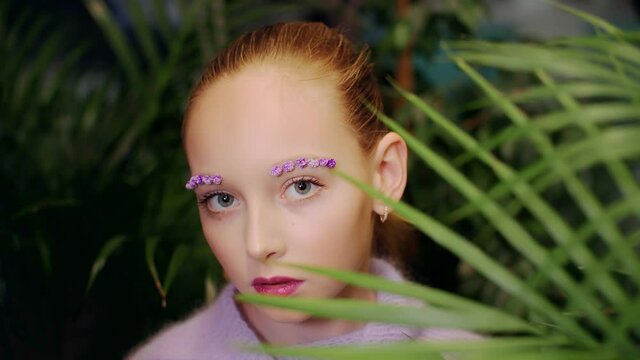 Close-up view of young girl face with beautiful makeup. Portrait of teenage fashion model with stylish vivid makeup looking at camera through green leaves