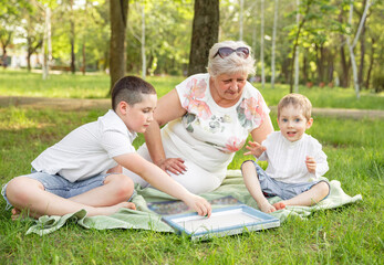 Naklejka na ściany i meble Cheerful extended family playing board game in their backyard. Family playing in the summer forest. Grandmother playing with her grandchildren at nature. Happy moments with grandmother outdoors.
