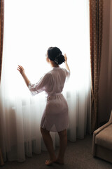 A girl in a dressing gown is standing near the window. A woman in a white lab coat. A girl with a beautiful figure in a dressing gown. A female silhouette in front of the window.