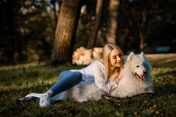 beautiful woman in white shirt is hugging her white dog samoyed outdoors in the park and sitting on the grass.
