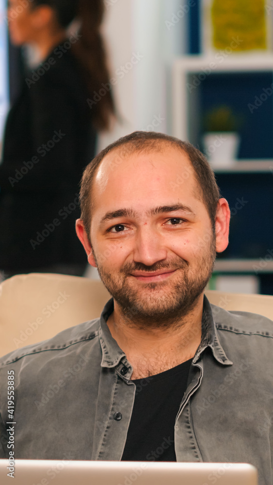 Poster manager sitting on couch in front of camera smiling while multiethnic team working in background wit