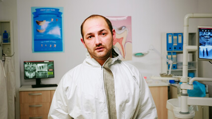 Obraz premium Portrait of exhausted stomatologist man taking off face shield looking at camera sitting in new normal dental office. pediatric nurse talking with child patient in background.