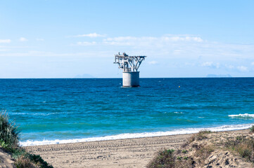 An old abandoned engineering structure with a cylindrical base is in the sea, a sandy beach with grass bushes in the foreground and a blue sky with light clouds in the background