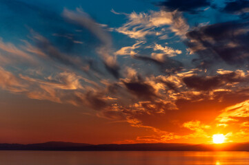 Fiery bright sunset over the sea and coastline with feather-like clouds against the blue sky