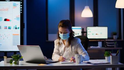 Manager woman with face mask working overtime in new normal business office checking team project,...