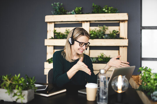 pretty, young, blond woman with stylish, modern black glasses sits in a sustainable office and has an online web meeting with a headset and is happy
