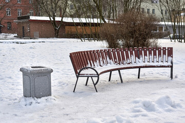 Trash can next to a bench covered with snow in a city park