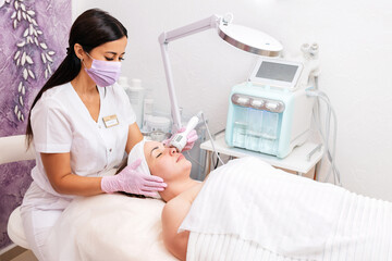 New normal. A cosmetologist in rubber gloves and medical mask does a rf-lifting procedure on the face. In the background, the Rf cosmetology device. Concept of professional cosmetology and health