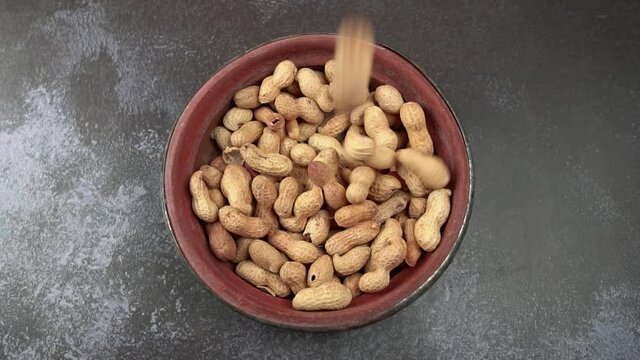 Top down view of peanuts falling into a rustic bowl on top of a dark grey background with copy space. Slow motion flay lay video of monkey nuts dropping into serving dish. 