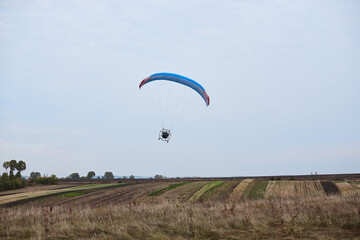 Paratrike with blue parachute flying high in the cloudy sky on autumn morning. Paragliding activity in rural countryside. Weekend leisure time and hobby. Extreme sport flight on paramotor machine.