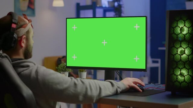 Gamer playing video games on powerful computer with green screen chroma key desktop mock-up display in gaming home studio. Player using RGB keyboard with isolated monitor streaming game wearing